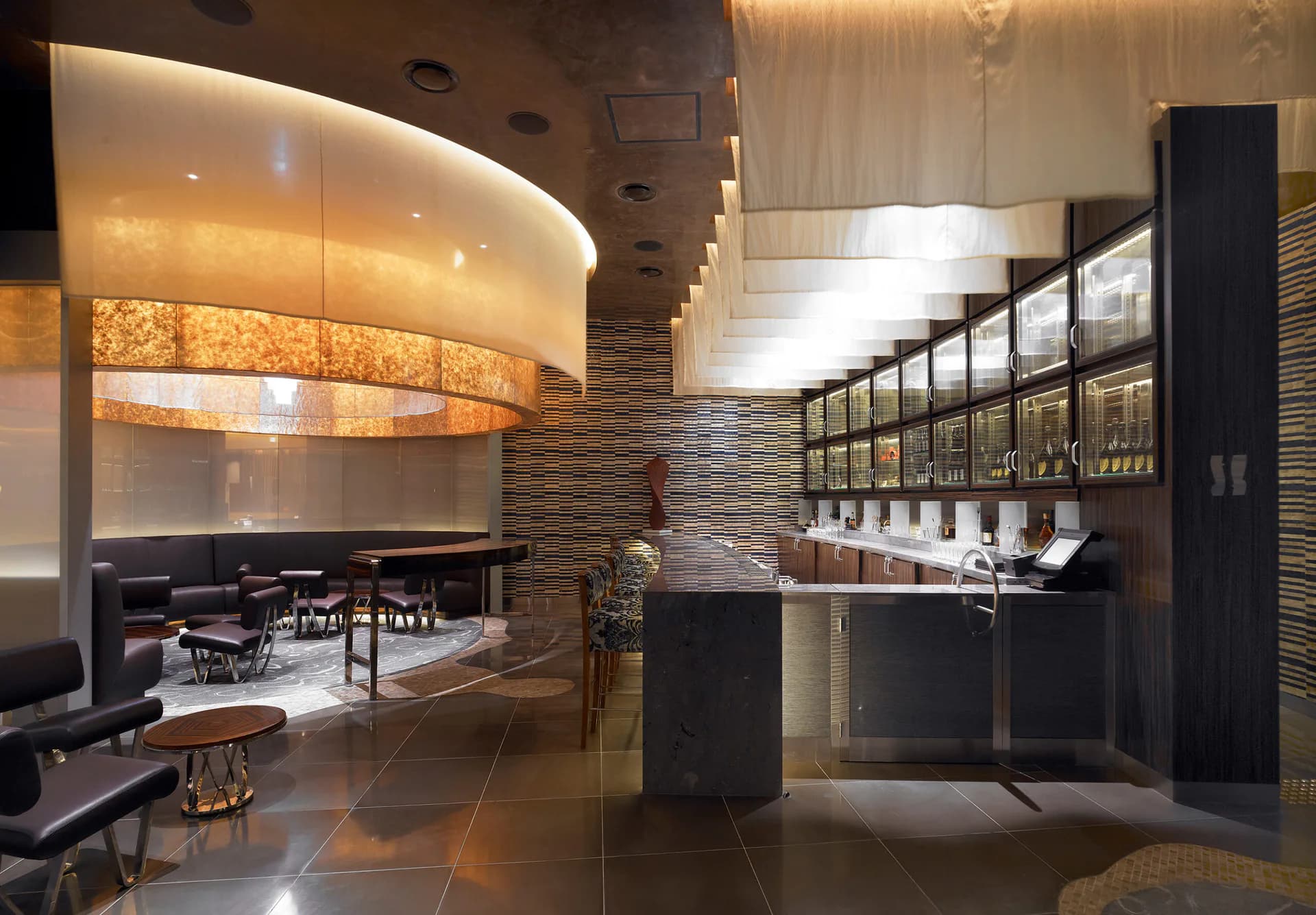 Modern luxury lounge with curved glass partitions, warm ambient lighting, dark textured walls, and sleek bar cabinetry by Nancie Giardina.