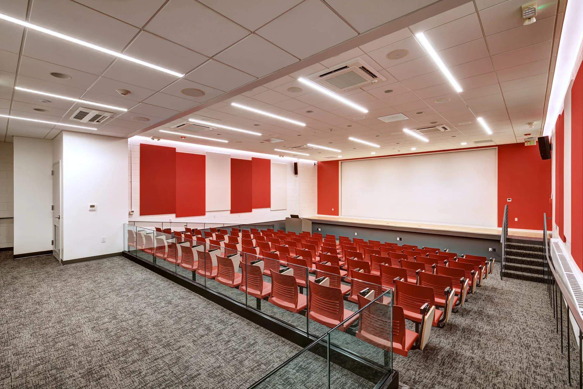 Modern auditorium with tiered red seats, white and red accent walls, linear ceiling lights, and a projection screen, designed by Nancie Giardina.