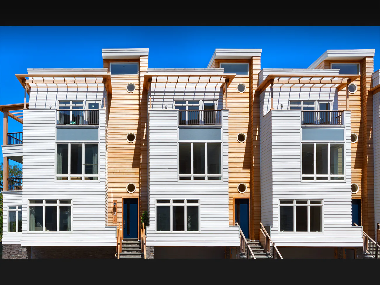 Three-story contemporary townhouses with white clapboard siding, wood accent panels, large multi-pane windows, and rooftop decks by Nancy J. Ruddy.