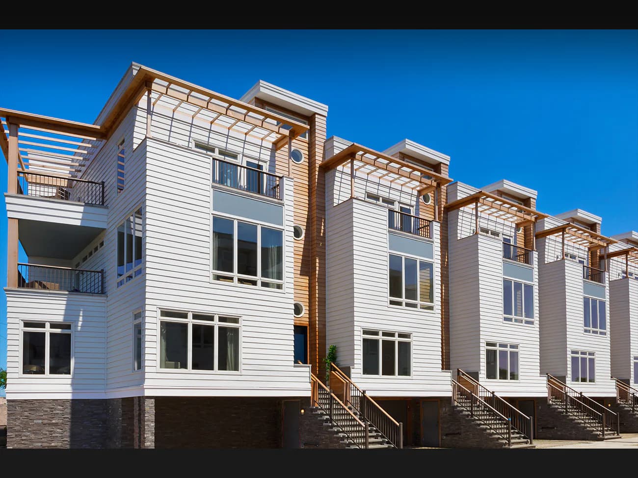 Row of contemporary residential townhouses with white siding, metal railings, and elevated bases on stone foundations against a clear blue sky by Nancy J. Ruddy.