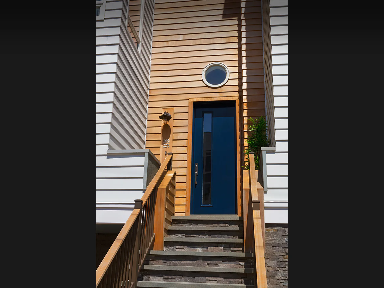 Residential entryway with wooden horizontal siding, blue door, round window, wooden railings, and brick steps by Nancy J. Ruddy.