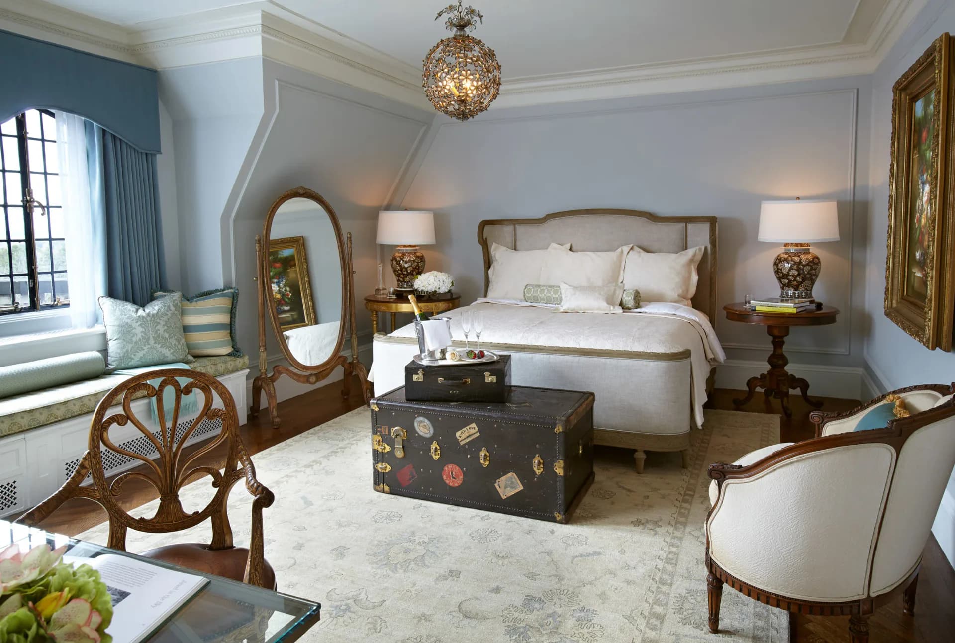 Elegant primary bedroom featuring a wood-framed upholstered bed, decorative trunk at foot, paired nightstands with brass mirrors, and window seating by Nancy Melius.
