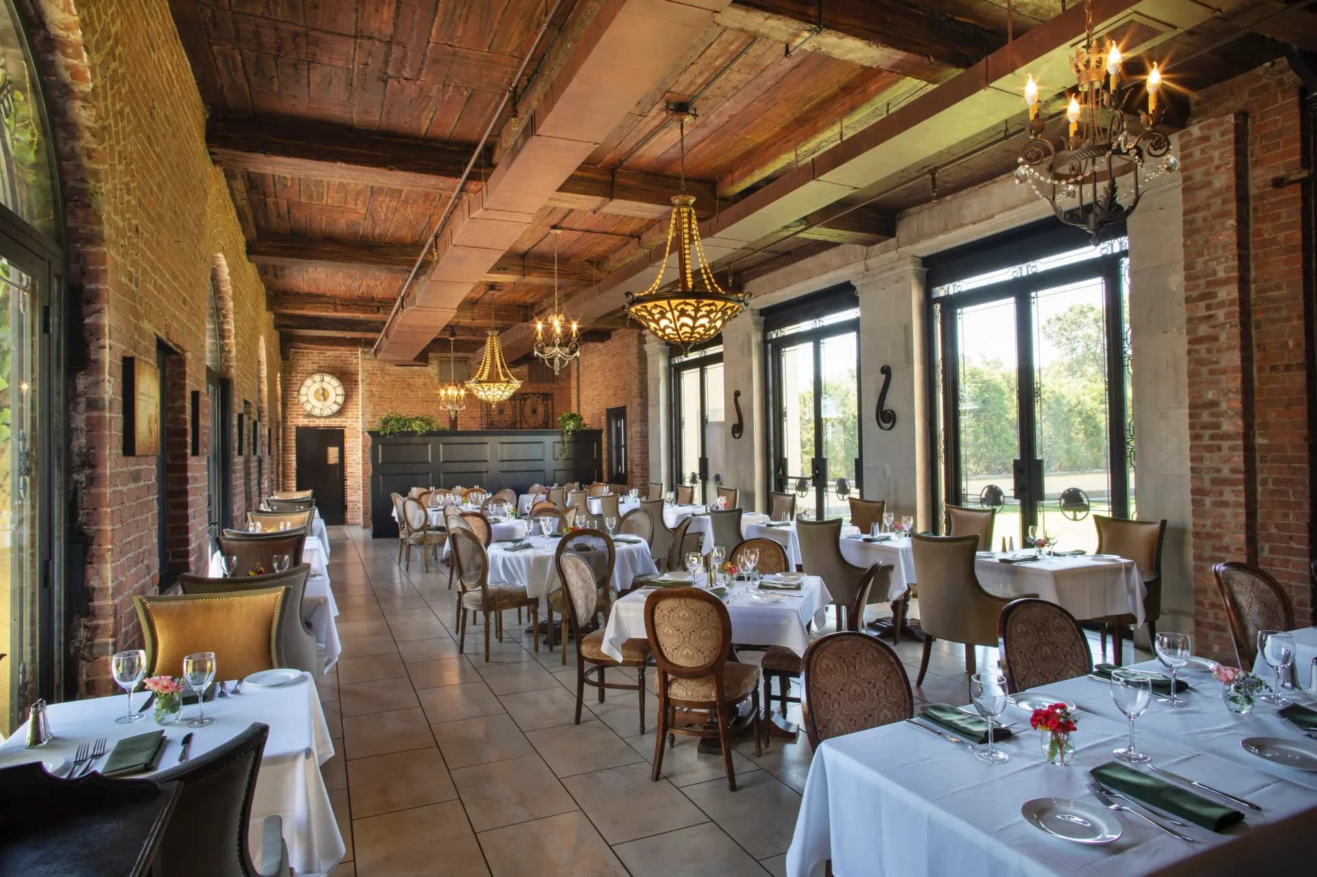 Elegant fine dining restaurant interior with exposed brick walls, wood beam ceiling, brass chandeliers, and white tablecloth-dressed tables arranged throughout the warm, sophisticated space by Nancy Melius.