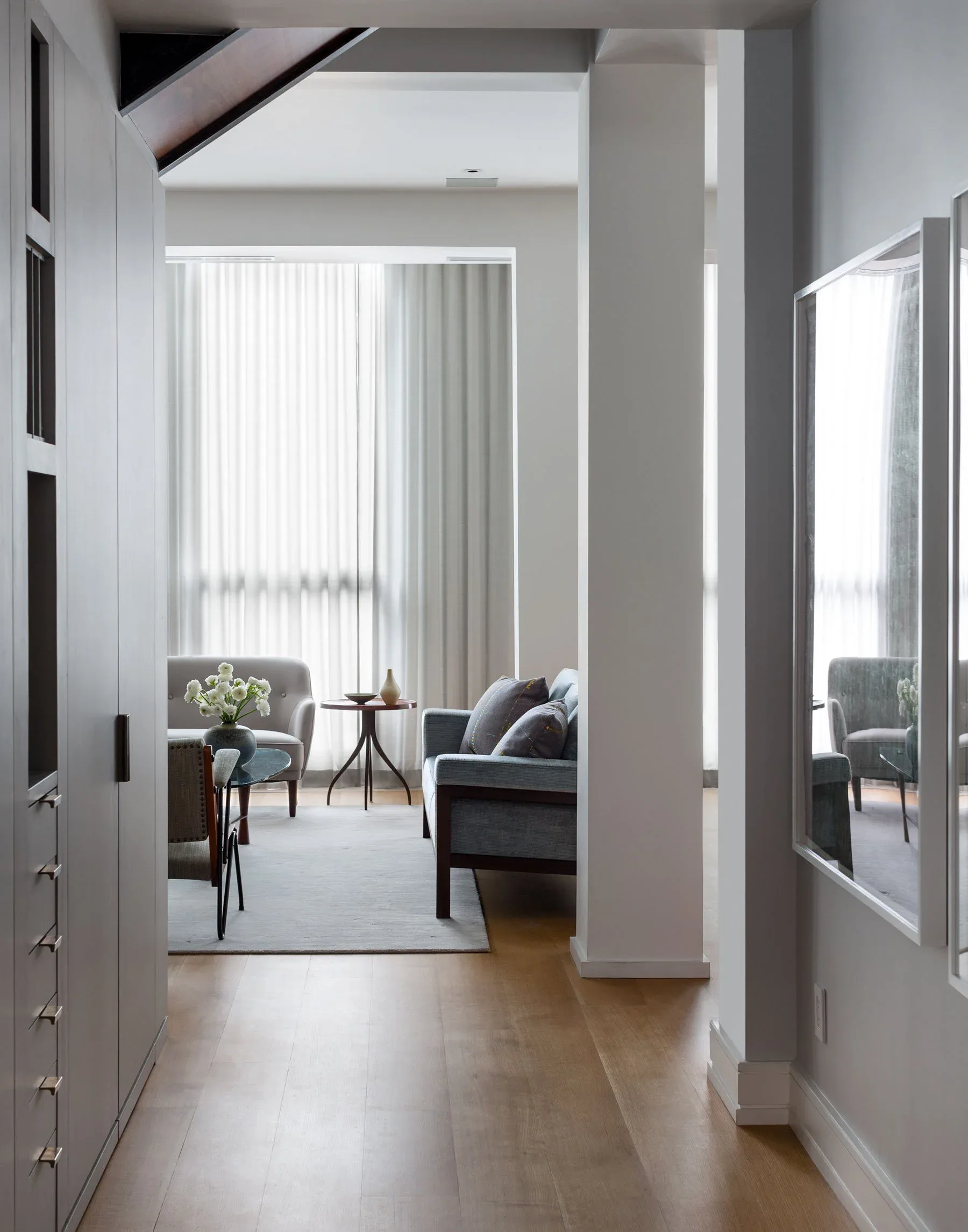 Modern residential hallway with light wood flooring leading to a bright living area with vertical blinds, upholstered seating, and minimalist furnishings by Nancy Nienberg.