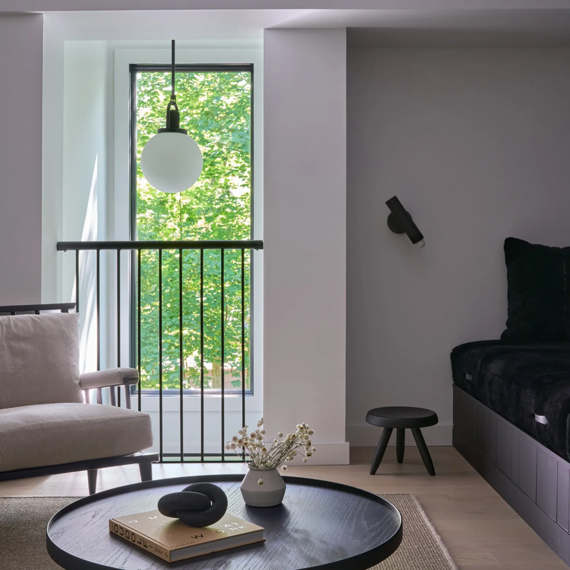 Modern living room with round marble table, black pendant light, floor-to-ceiling windows overlooking greenery, and minimalist furnishings by Nancy Nienberg.