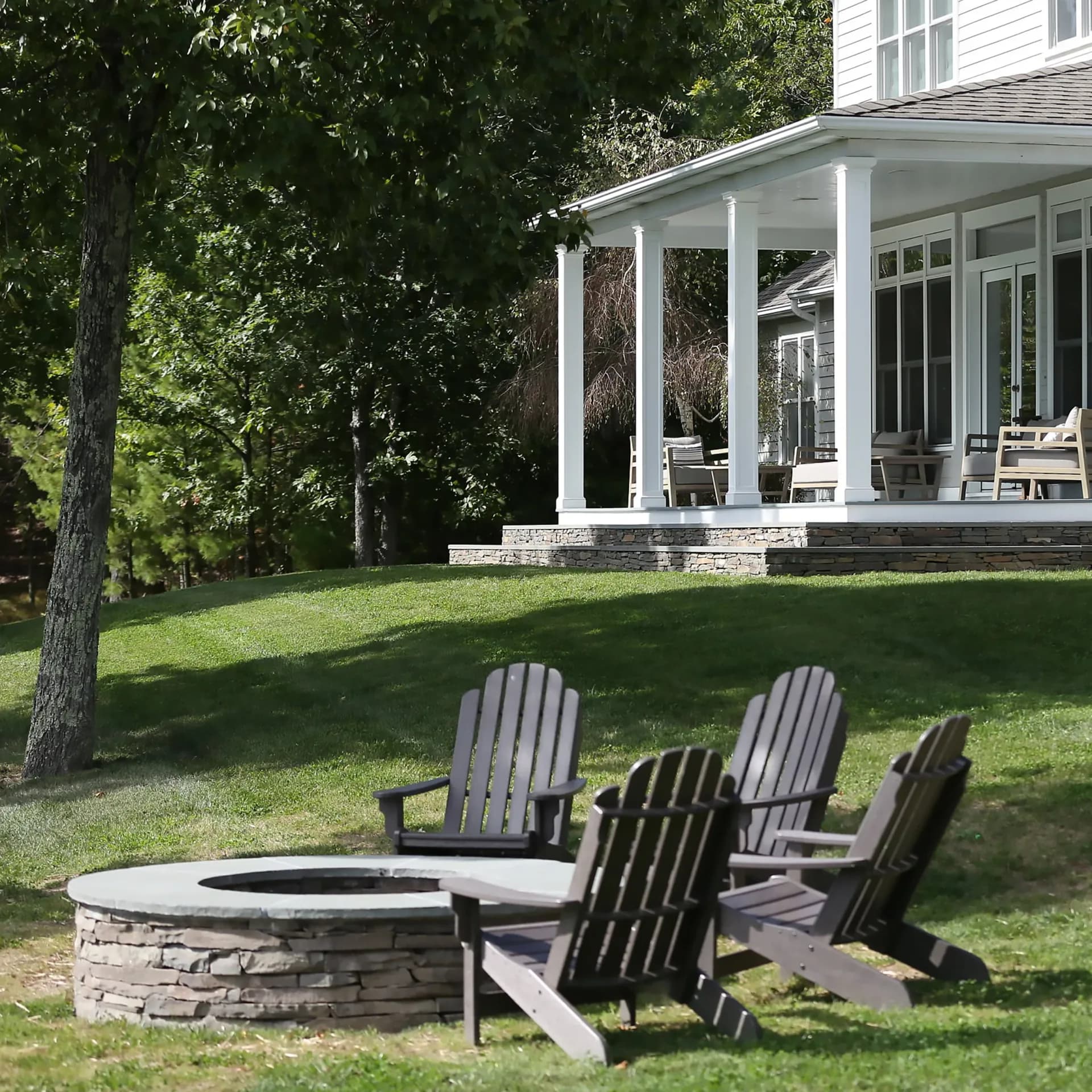 Residential backyard with fire pit seating area, Adirondack chairs, manicured lawn, and modern white porched house by Nancy Nienberg.