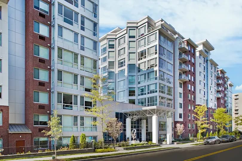 Modern multi-story residential building with mixed materials including brick and glass facade, ground-level columned entrance, and landscaped streetscape by Nancy Zito.
