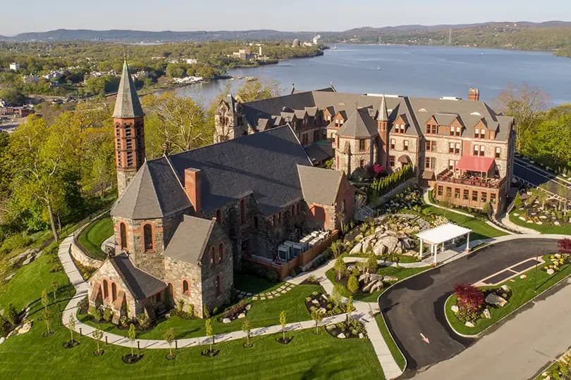 Aerial view of a brick Gothic Revival church and adjacent Victorian mansion overlooking a lake, with manicured gardens and landscaping by Nancy Zito.
