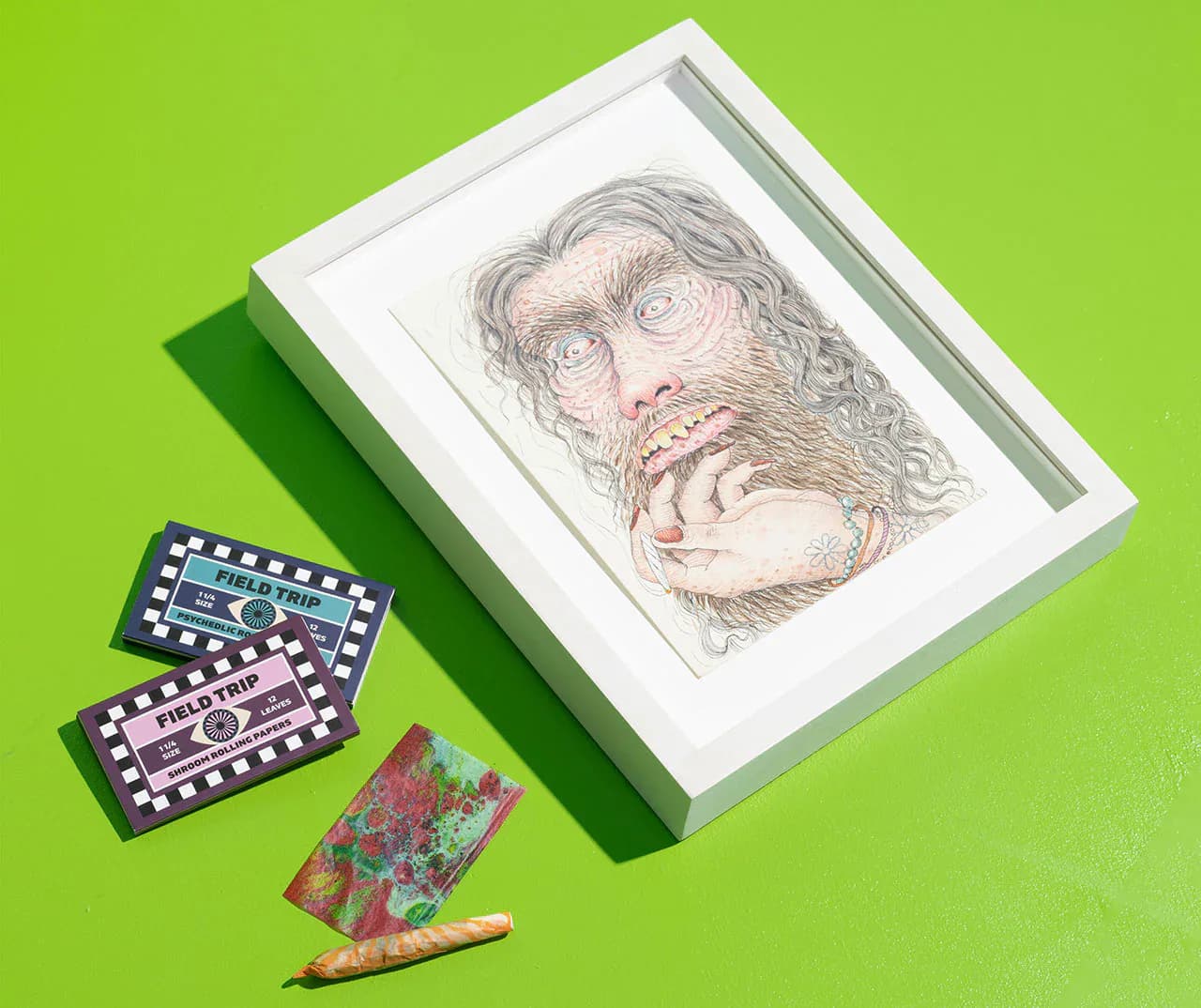 Styled flat lay composition featuring a framed portrait drawing, decorative patterned items, and colored pencil on bright green background by Naomi Clark.