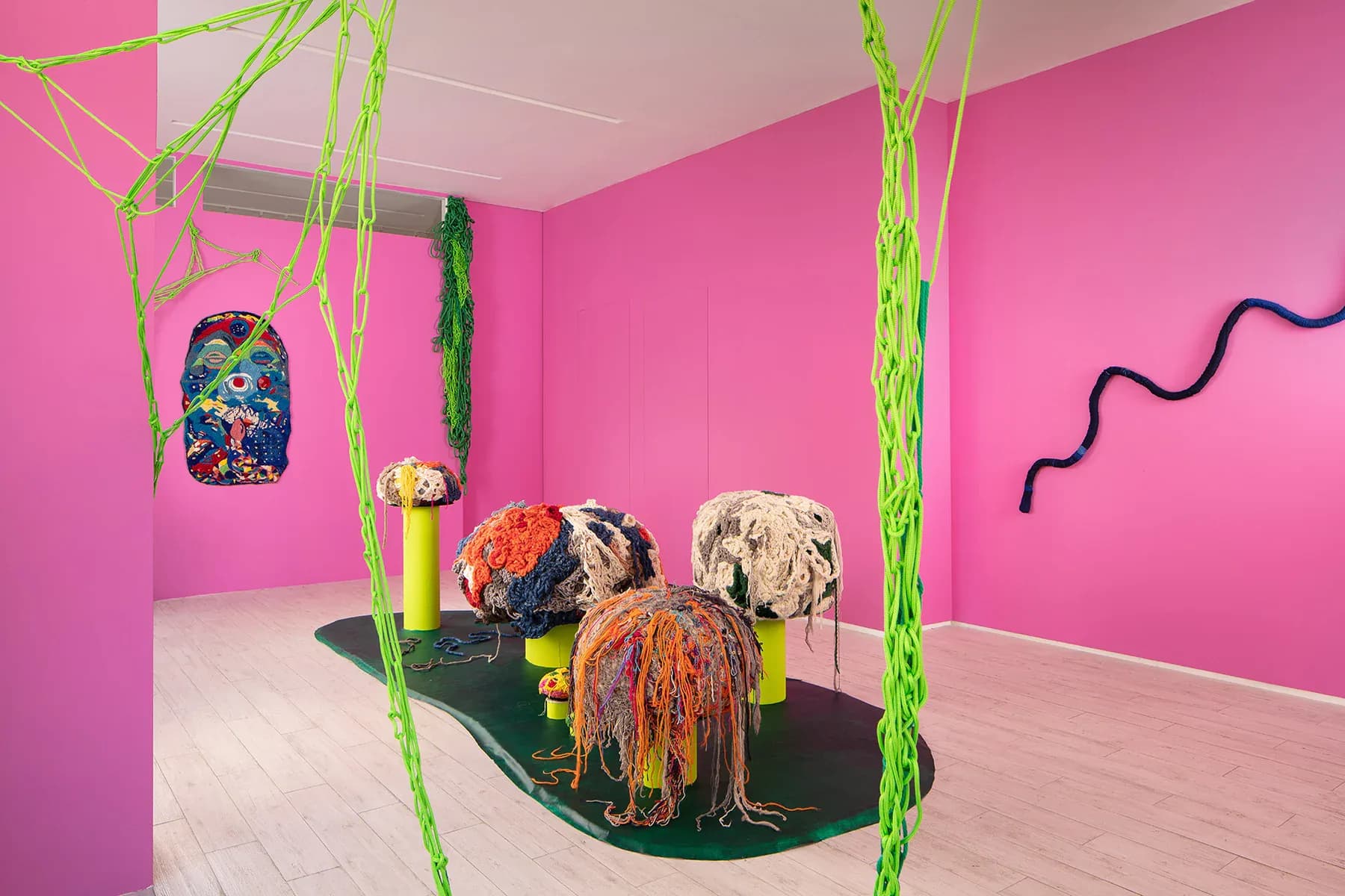 Vibrant pink gallery installation featuring hanging neon-green sculptural elements, colorful fiber artworks on black platform, and curved wall details by Naomi Clark.