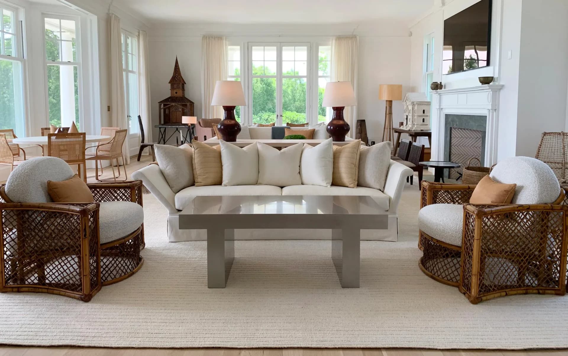 Elegant living room with cream sectional sofa, woven armchairs, concrete coffee table, fireplace, and large windows overlooking gardens by Naomi Stroh.