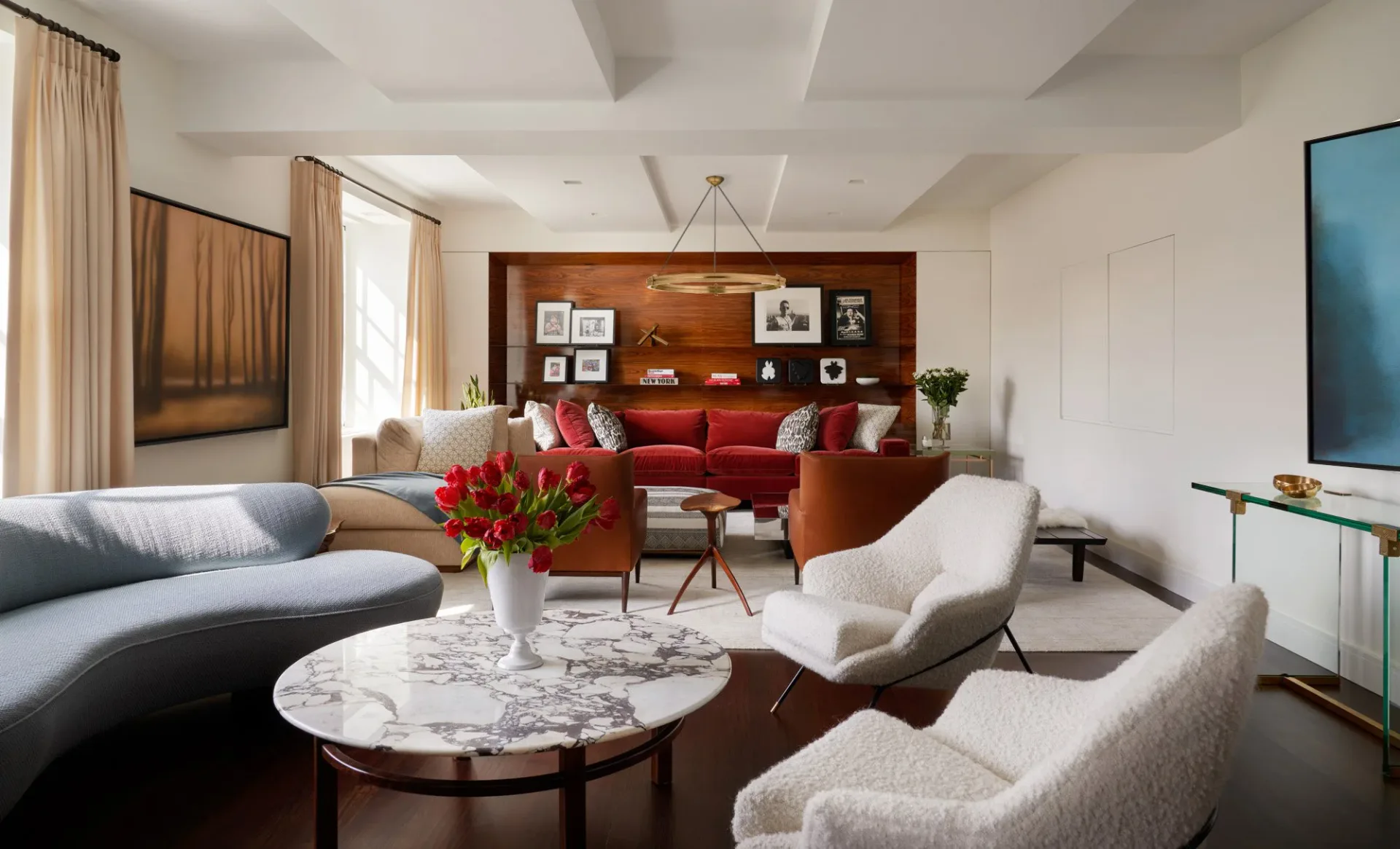 Contemporary living room with curved gray sofa, red accent seating, marble coffee table, and warm wood shelving, designed by Naomi Stroh.