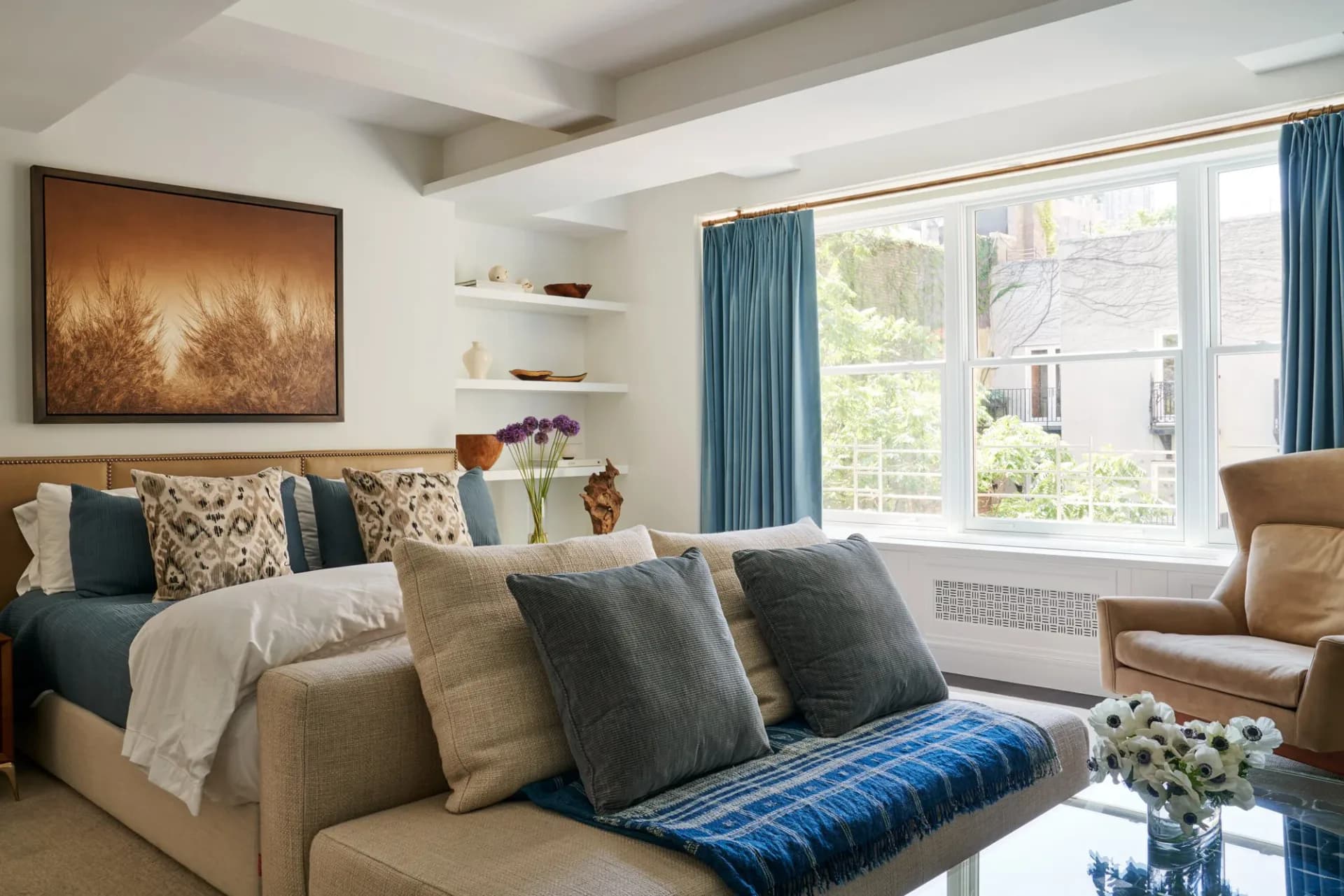 Luxurious master bedroom with teal curtains, upholstered bed with patterned pillows, floating shelves, and seating area featuring warm wood and neutral tones by Naomi Stroh.
