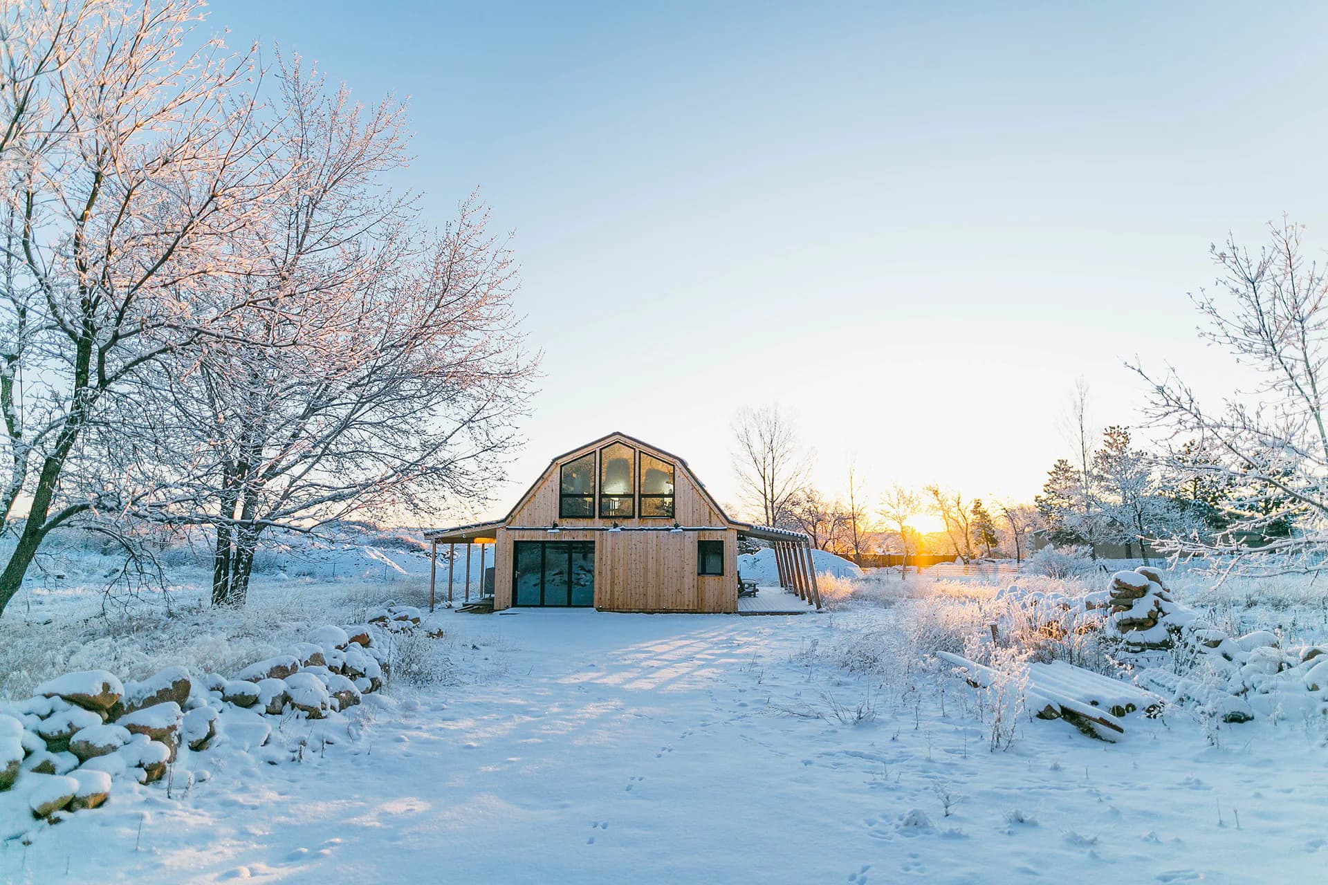 A modern barn-style house with wood and metal exterior sits in a snowy winter landscape at sunset, surrounded by bare trees and frost-covered fields, designed by Nasim Mossabeh.