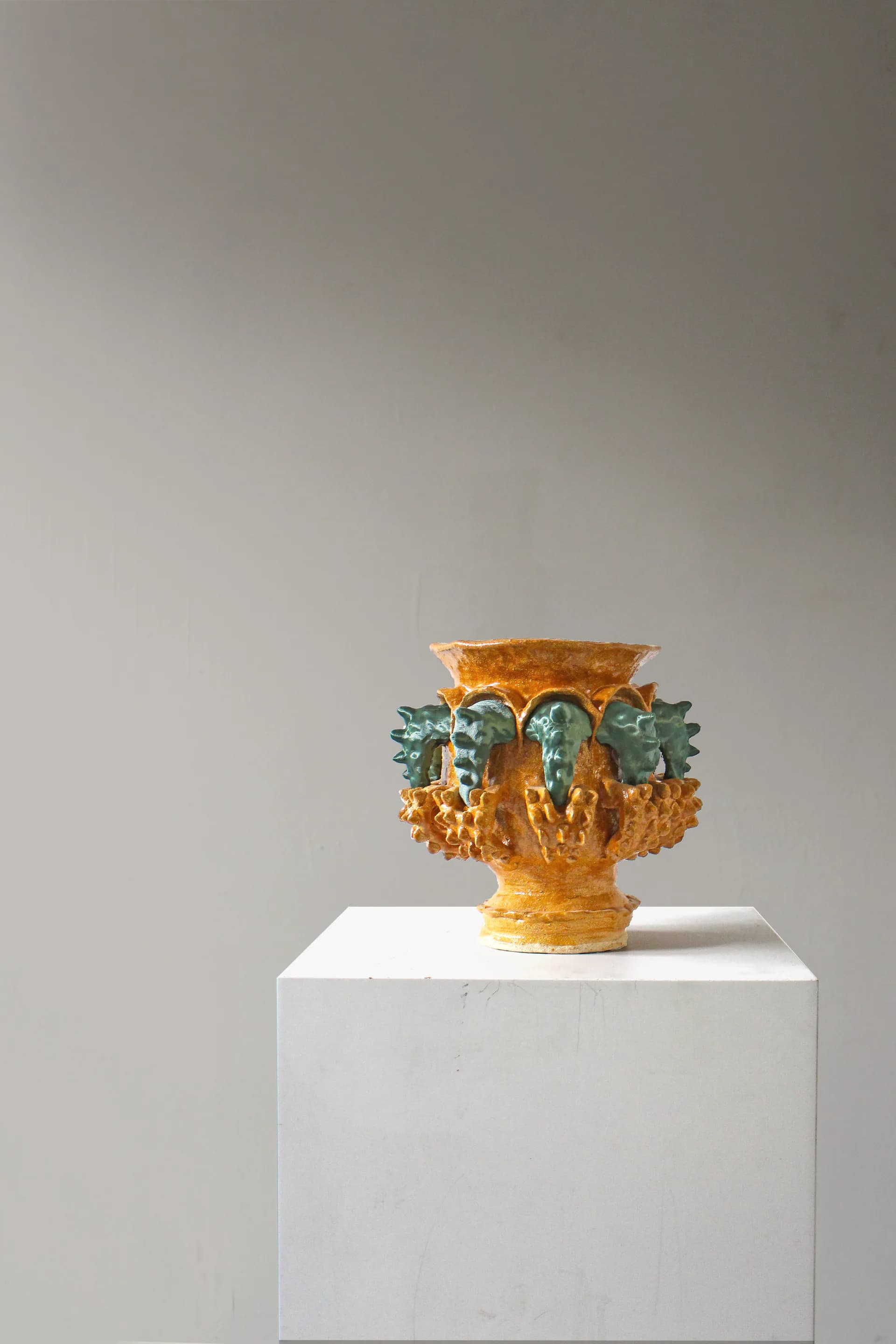 Ornamental vessel with golden textured body and green leafy handles displayed on white plinth against neutral background by Natalia Triantafylli.