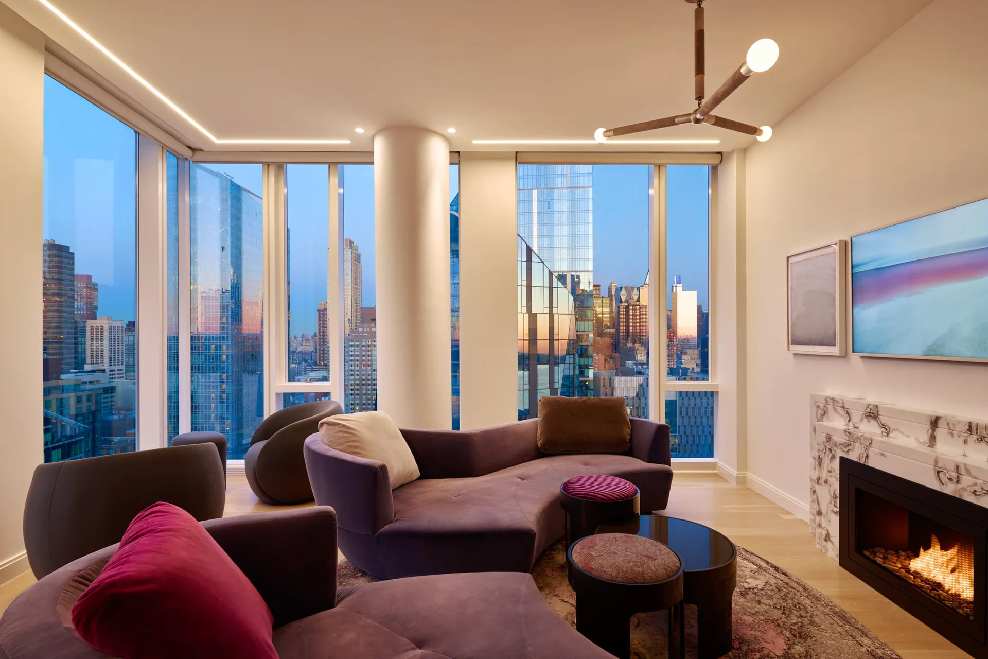 High-rise living room with floor-to-ceiling windows overlooking Manhattan skyline, featuring plush purple and brown seating, central cylindrical column, fireplace, and modern lighting by Natalia Tutka.