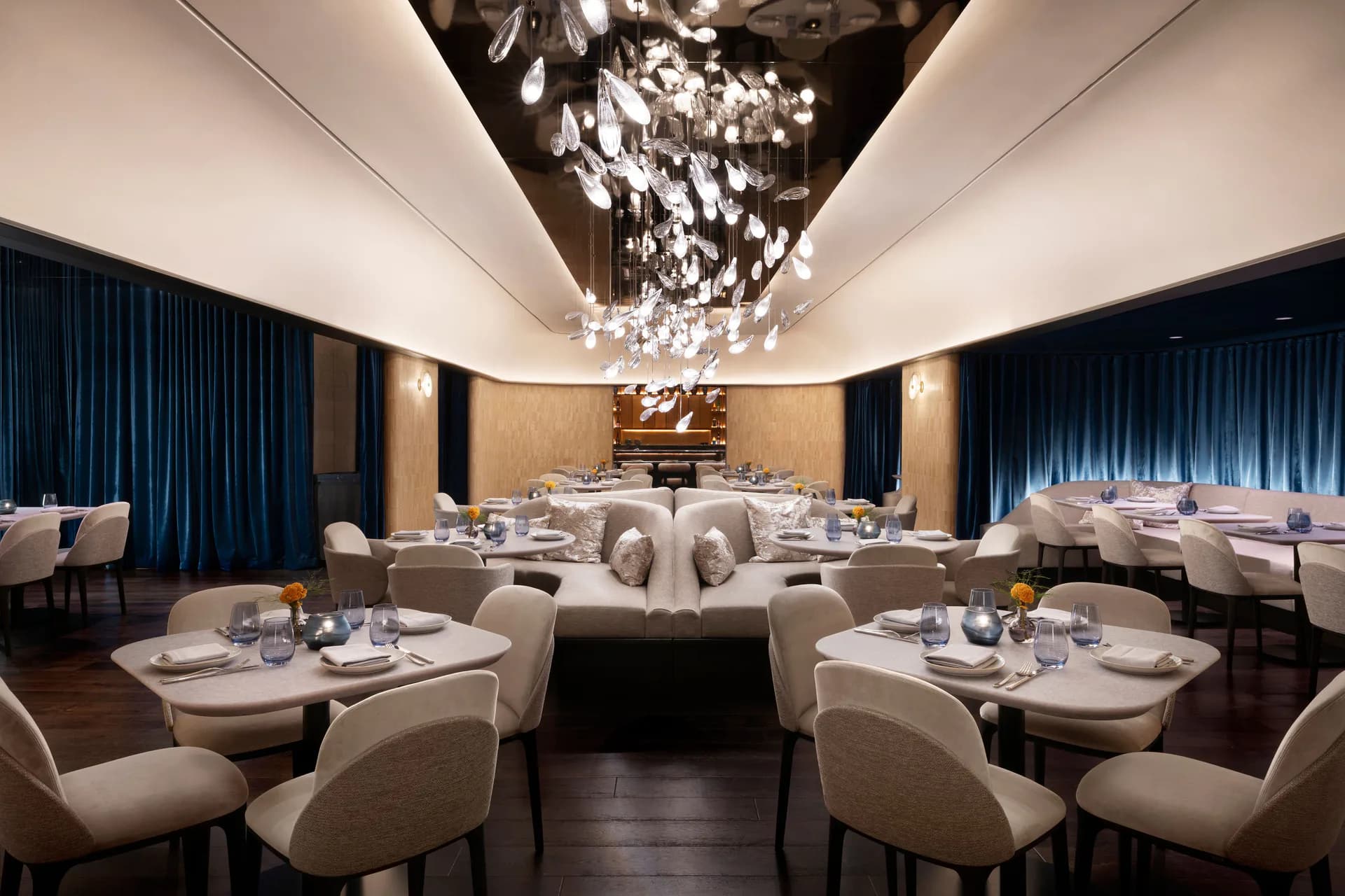 Luxurious restaurant dining room with symmetrical layout, cream-colored curved chairs, round tables, deep blue curtains, ornate ceiling fixture, and warm golden accent lighting by Natalie Imran.