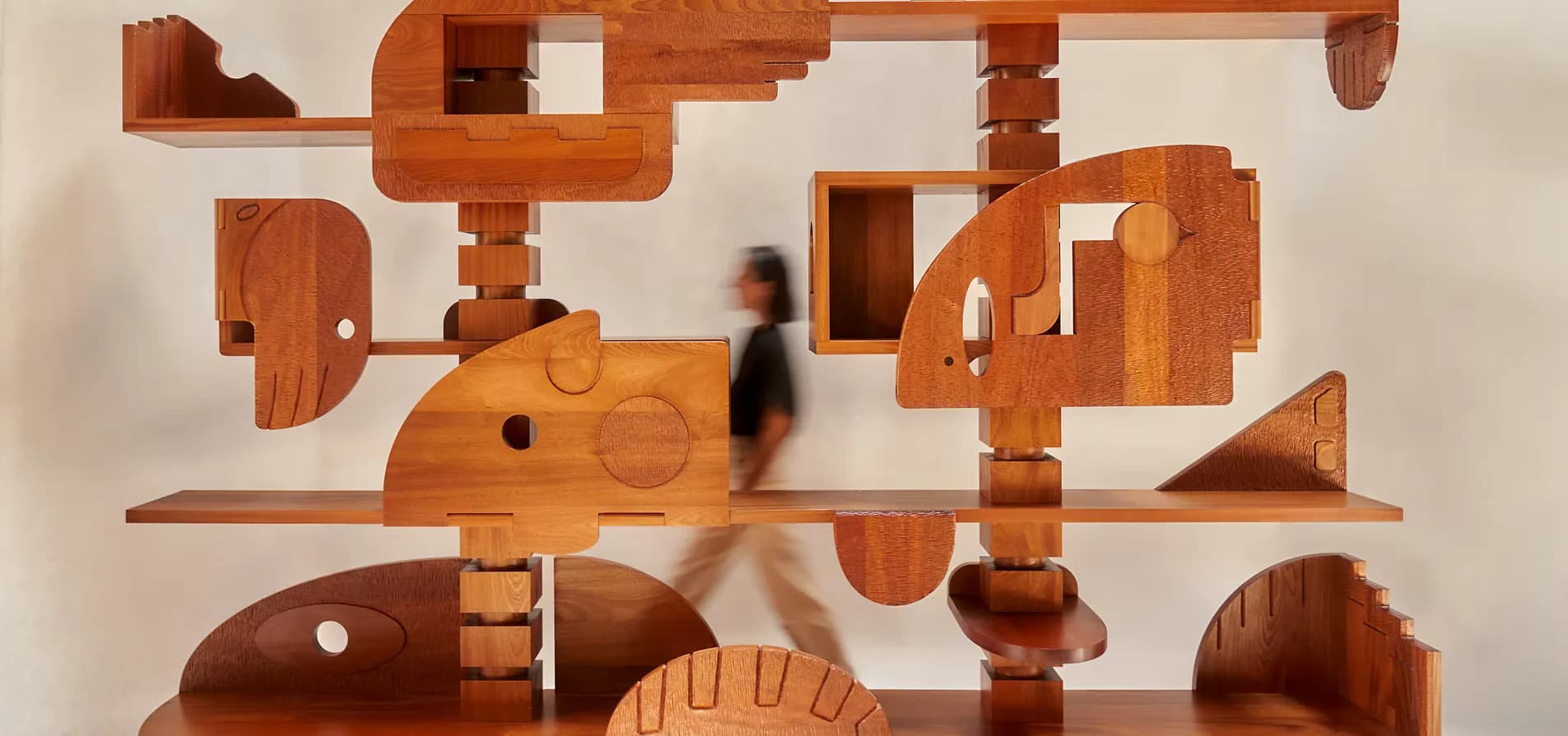Wooden wall-mounted sculptural installation with interlocking geometric forms in warm cherry tones, featuring a person walking past for scale, by Natalie Shook.