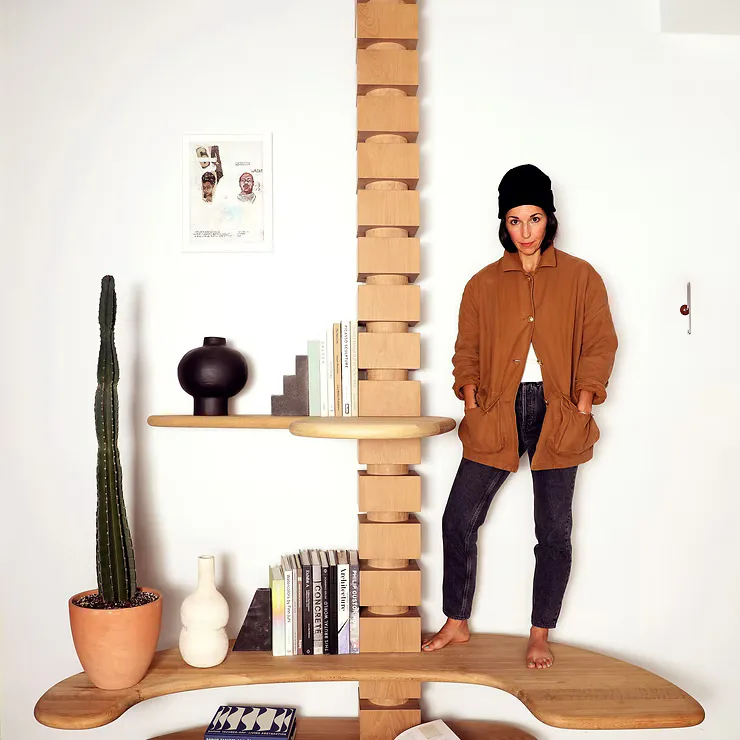 Wooden shelving installation with stacked box-frame design holding books, plants, and decorative objects, featuring curved wooden base platform by Natalie Shook.