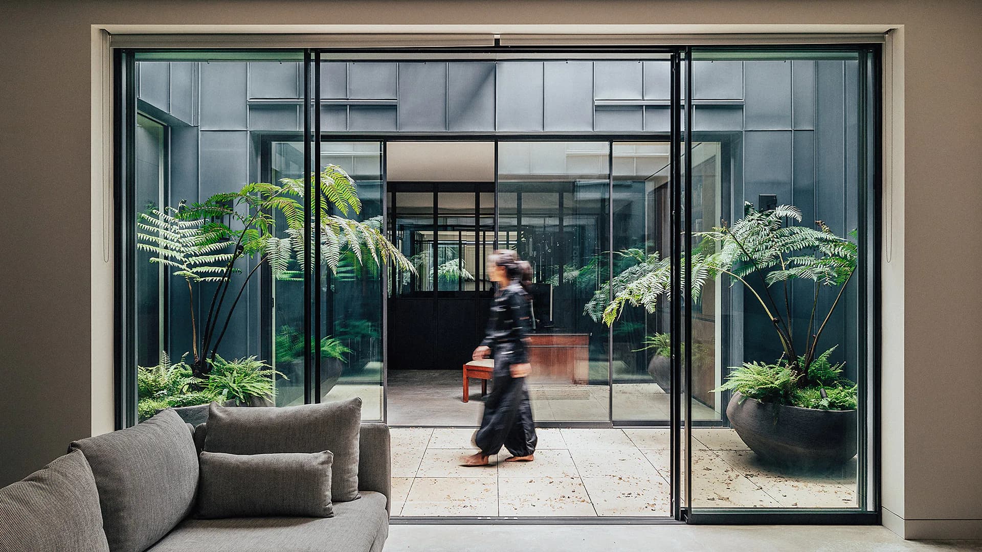 Modern interior with gray seating opens onto a courtyard through black-framed glass doors, flanked by potted plants and greenery, by Naushin Rahman.