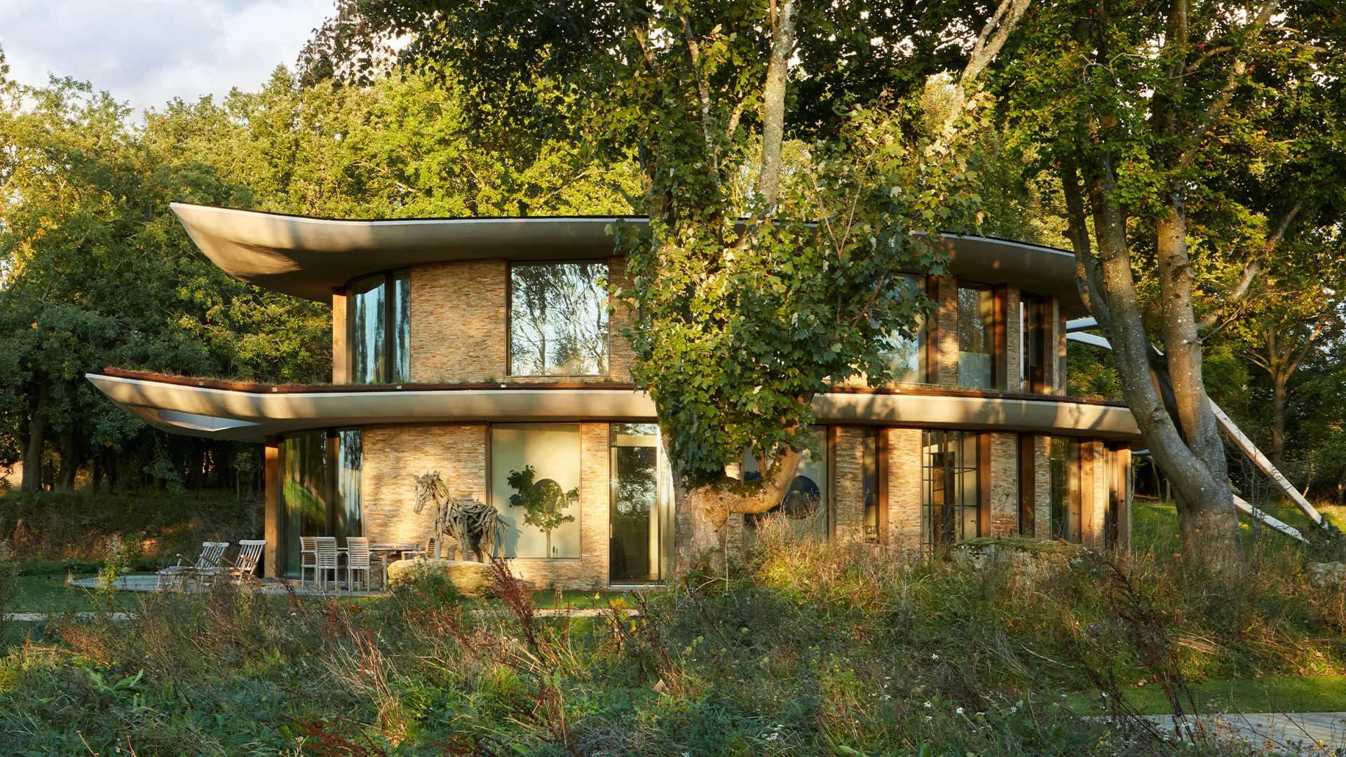 Modern residential structure with curved overhanging roofs nestled among mature trees, featuring stone and glass materials in warm afternoon light by Naushin Rahman.