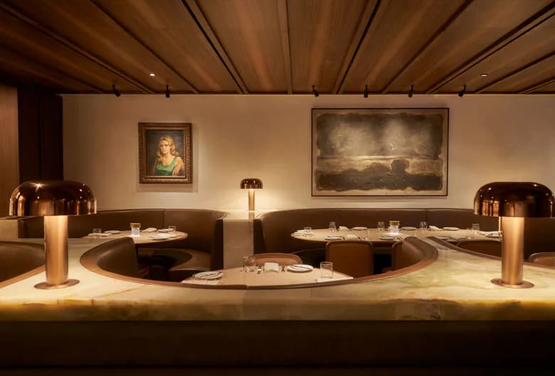 Luxurious restaurant interior with curved banquette seating, warm wood ceiling, dramatic artwork, and ambient bronze lighting by Naushin Rahman.