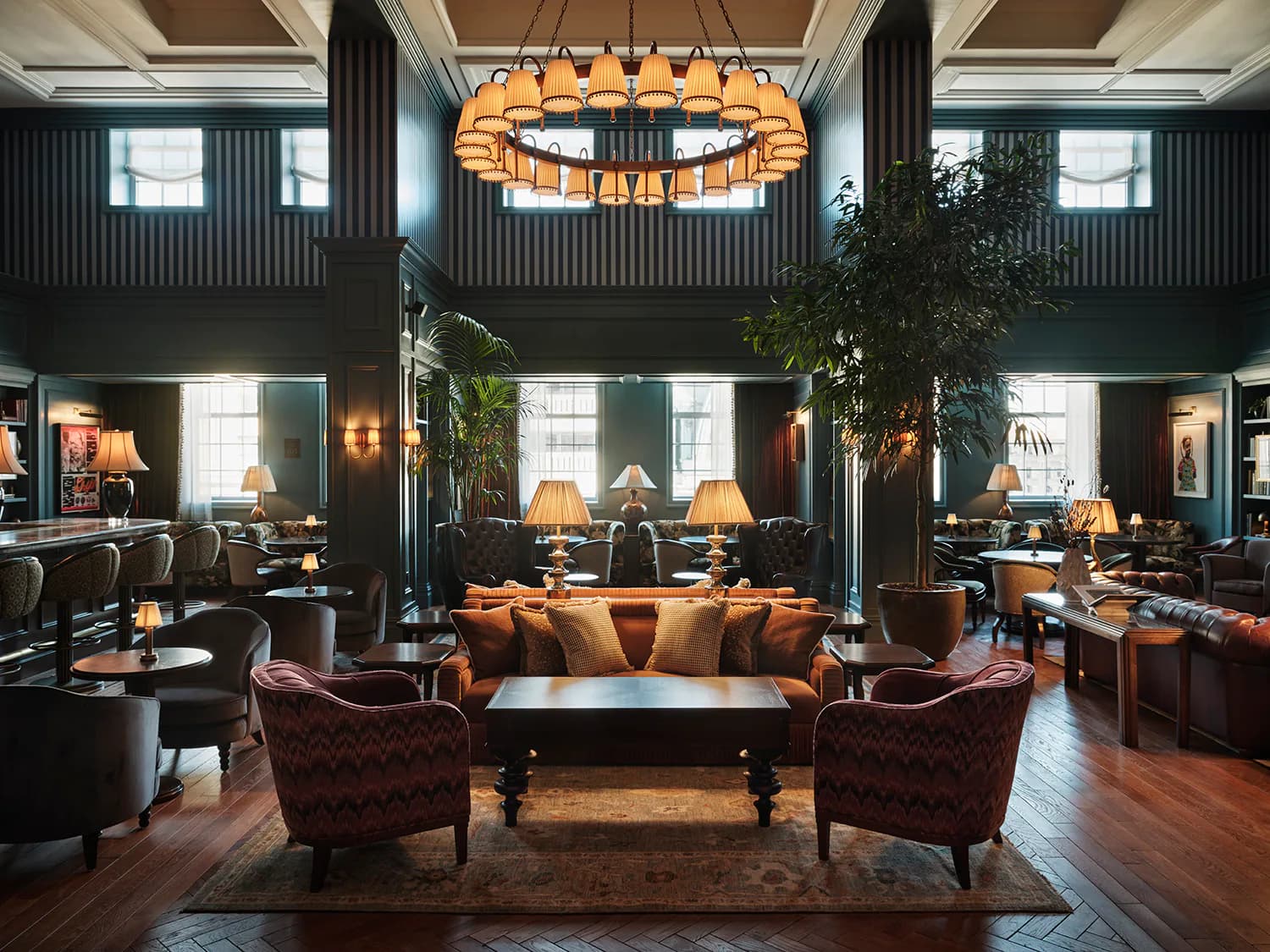 Luxurious hotel lobby with two-story ceilings, cognac leather sofa, burgundy accent chairs, warm brass lighting, dark green walls, and rich wood flooring by Nduka Aliberi.