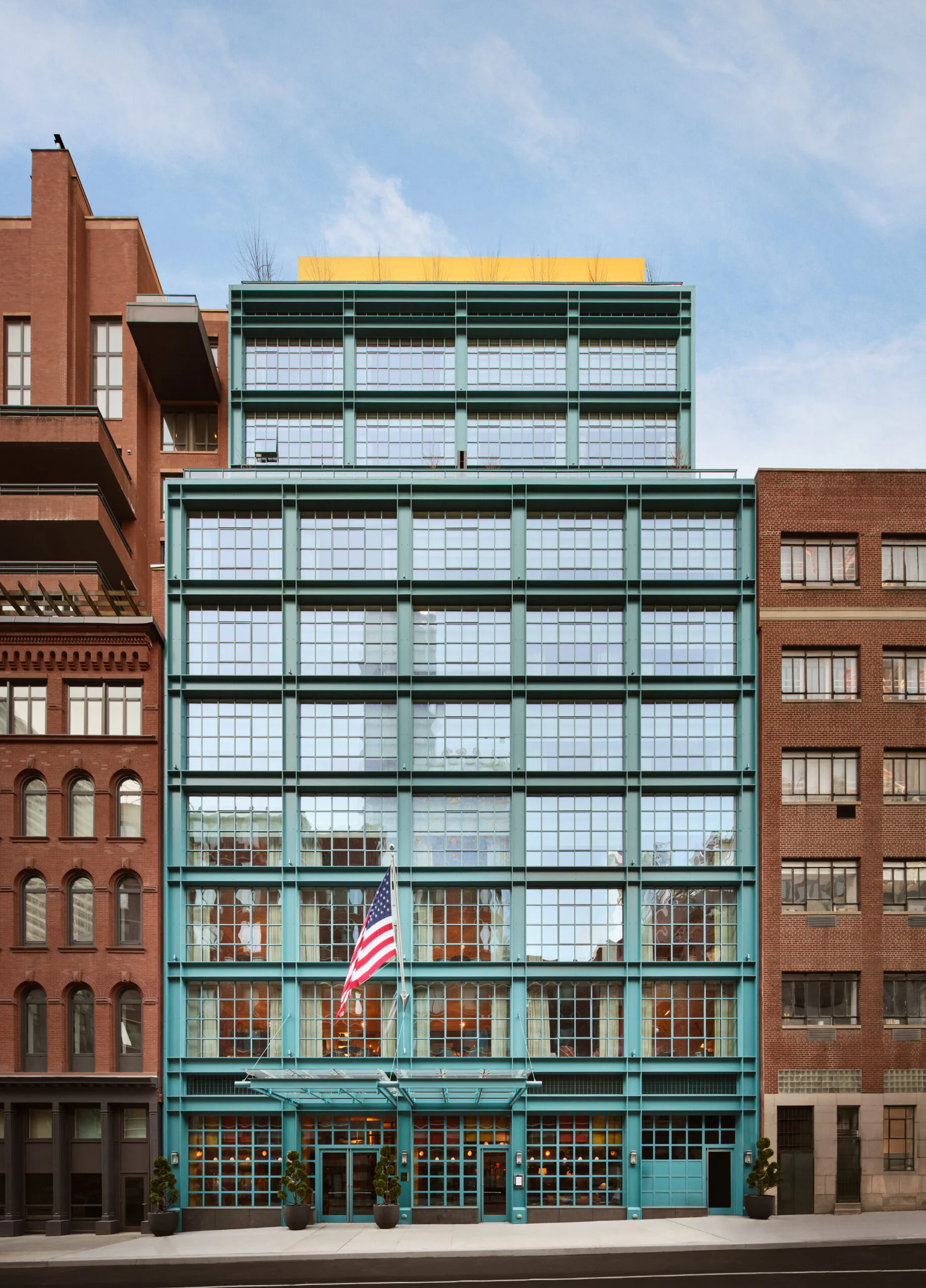 Striking turquoise and green facade with gridded glazing and yellow roof element, flanked by historic red brick buildings in urban setting by Nduka Aliberi.