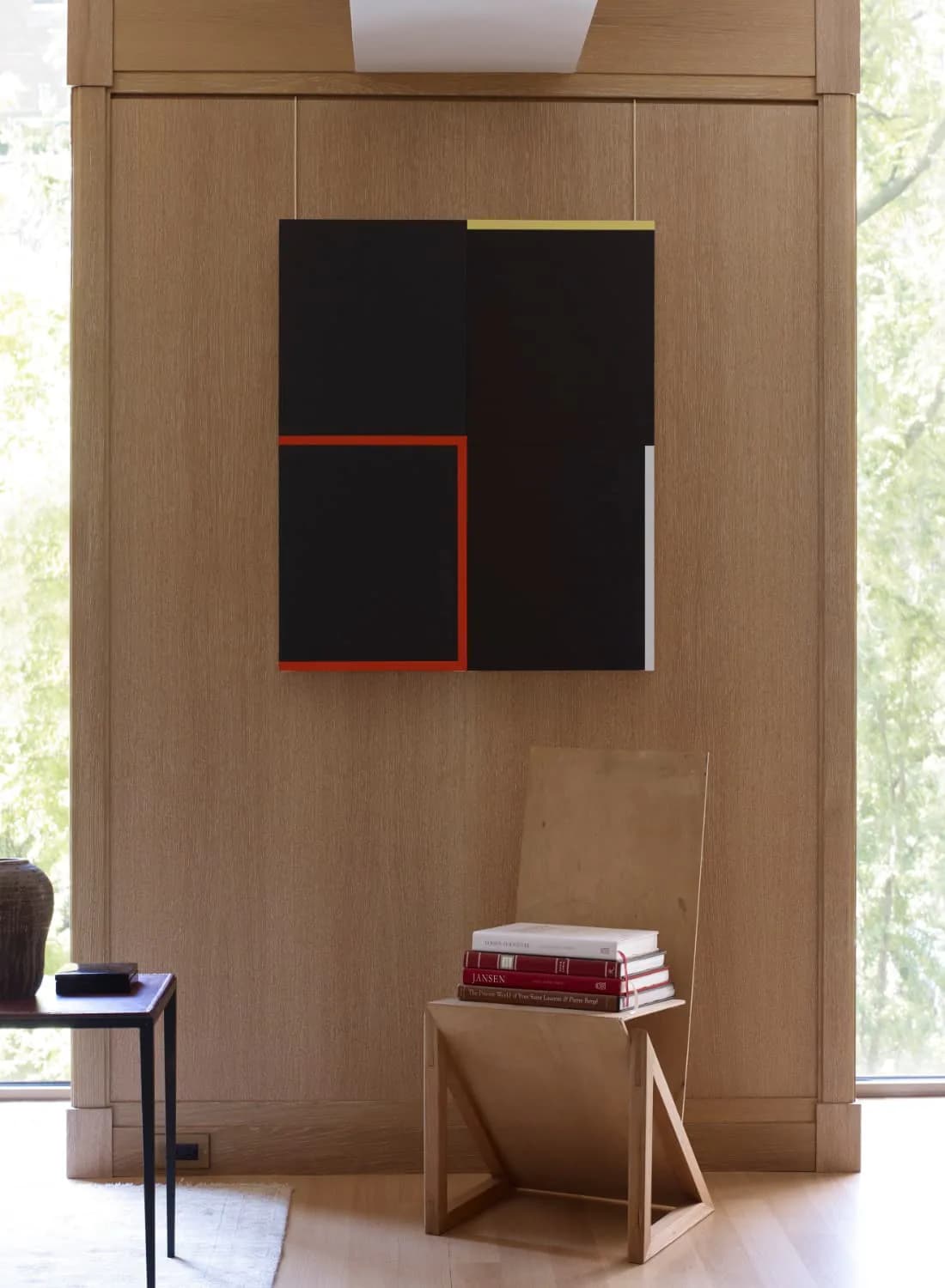 Modern interior with light wood paneling features a geometric abstract artwork with black, red, and gold elements above a minimalist wooden side table holding stacked books, designed by Neal Beckstedt.