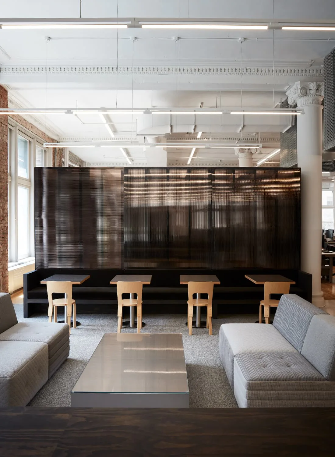 Modern loft interior with black metal-framed partition, light wood bar seating, neutral upholstered seating, and industrial ceiling details by Neal Beckstedt.