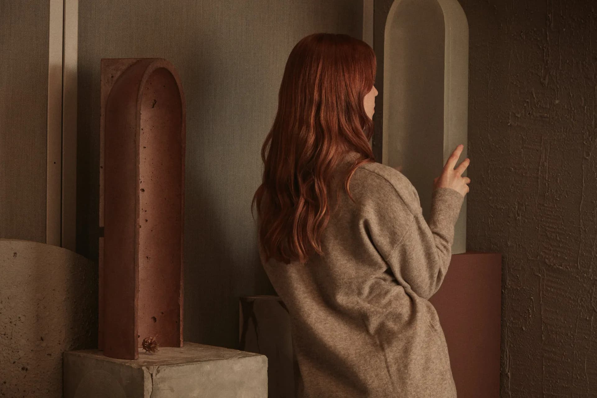 Warm minimalist interior with terracotta cylindrical sculptures and curved architectural elements, featuring a woman in beige cardigan, designed by Nel Verbeke.
