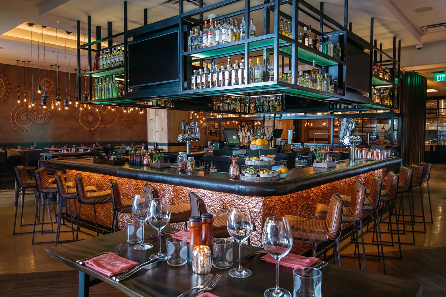 Modern bar interior featuring a two-level island with copper-textured facade, blue steel framework, extensive bottle display, and warm ambient lighting by Nelson Jenkins.