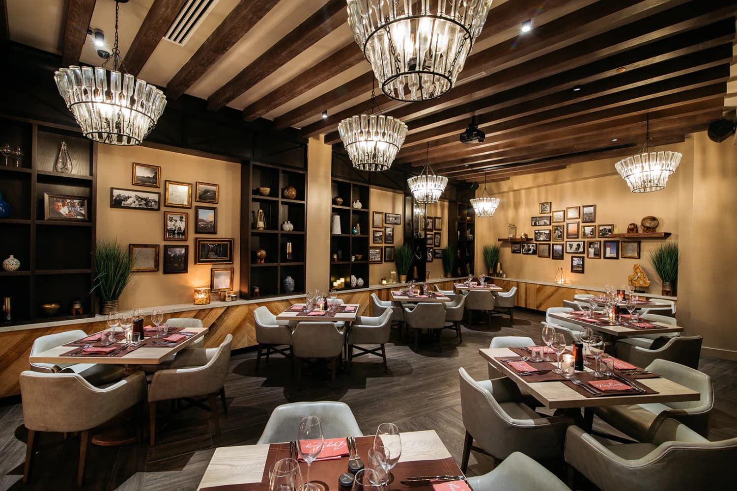 Elegant dining room with cream and black palette, exposed wood beam ceiling, crystal chandeliers, curved banquette seating, and gallery wall display by Nelson Jenkins.