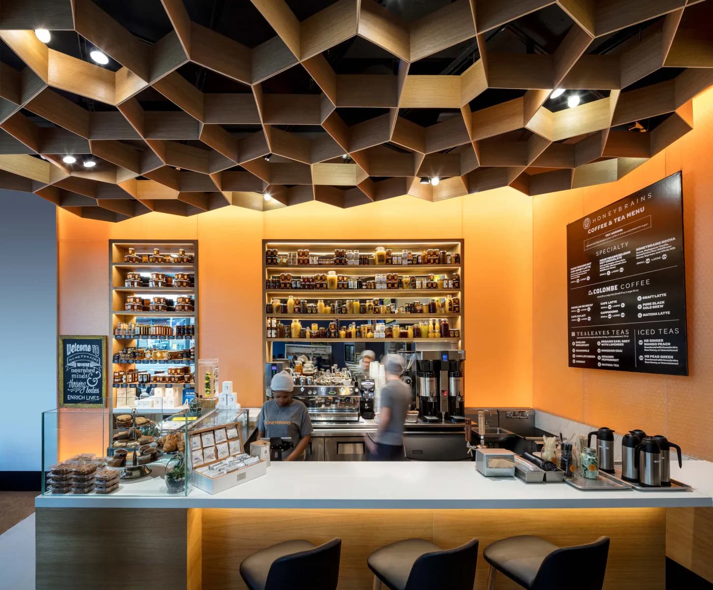 Modern coffee shop interior featuring warm orange walls, geometric ceiling panels with integrated lighting, white counter with black stools, and shelving displays, designed by Nelson Jenkins.