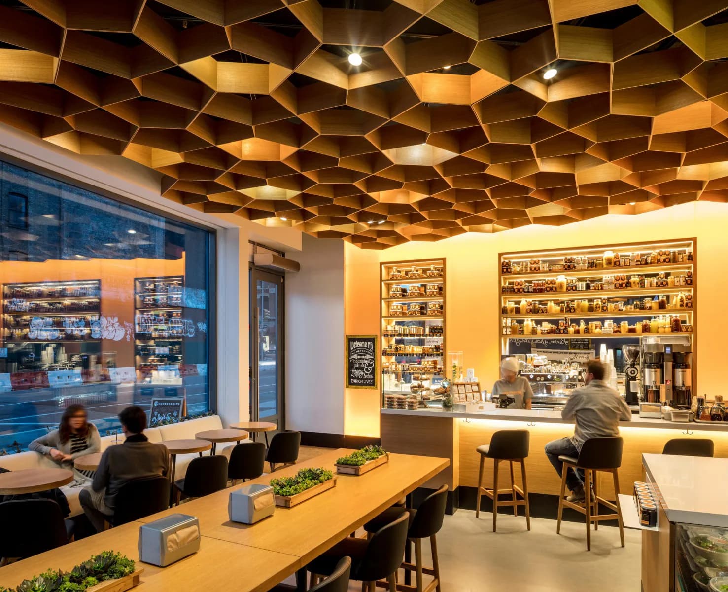 Modern retail interior featuring a geometric wood-panel ceiling with integrated lighting, yellow accent walls displaying product shelving, wooden tables with black stools, and blue storefront windows by Nelson Jenkins.
