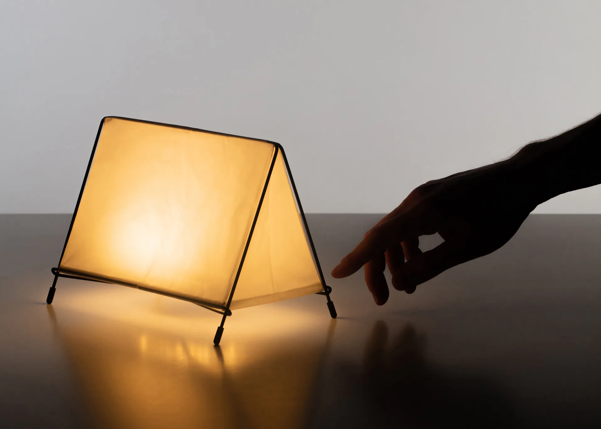 Minimalist table lamp with geometric trapezoid-shaped paper shade emitting warm golden light, photographed with hand gesture interaction by Nicholas Baker.