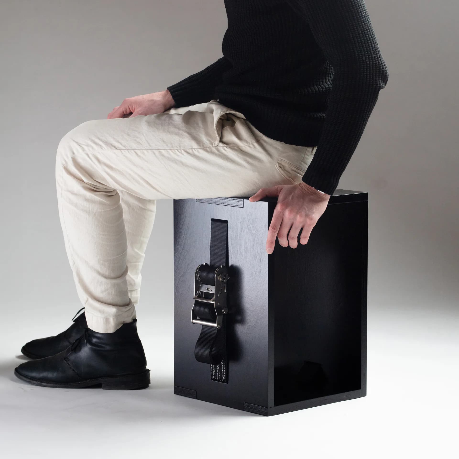 Product design photograph featuring a minimalist black cube with integrated leather straps, styled with a seated figure in cream and black clothing against a neutral background by Nicholas Baker.