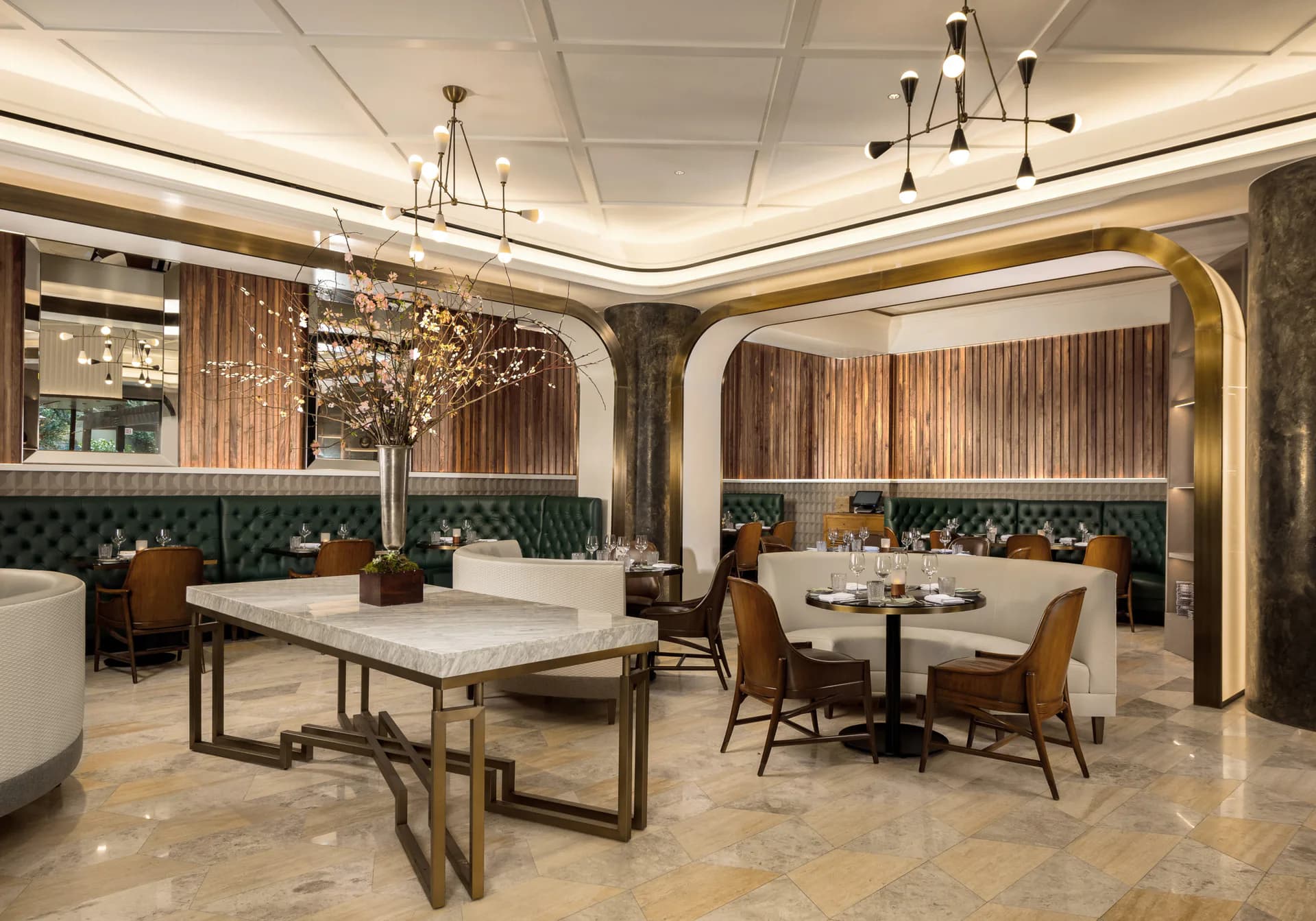 Luxurious restaurant interior with green velvet banquettes, marble flooring, brass-framed archways, and pendant lighting in warm tones by Nicholas Caruos.