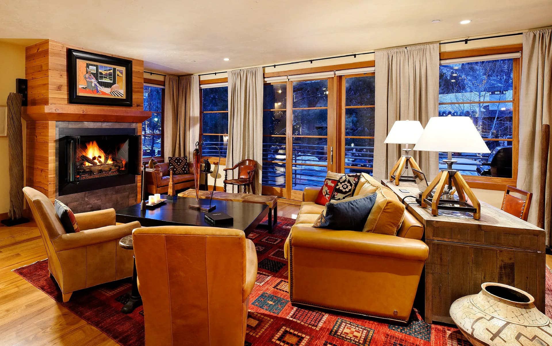 Luxury living room with warm cognac leather seating, wood fireplace, large windows overlooking snowy landscape, and red patterned rug by Nicholas Caruos.