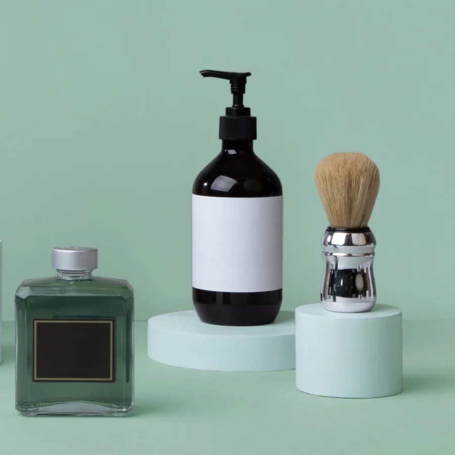 Styled product photography featuring a green glass perfume bottle, white and black soap dispenser with pump, and natural bristle shaving brush on pedestals against a soft sage green background by Nicholas Devlin.