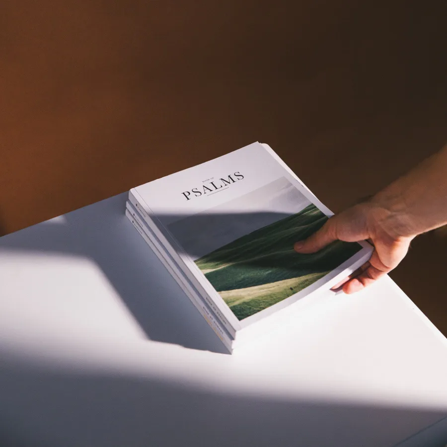 Hand holding open a white book titled "PSALMS" with green landscape imagery visible inside, photographed in natural sunlight by Nicholas Devlin.