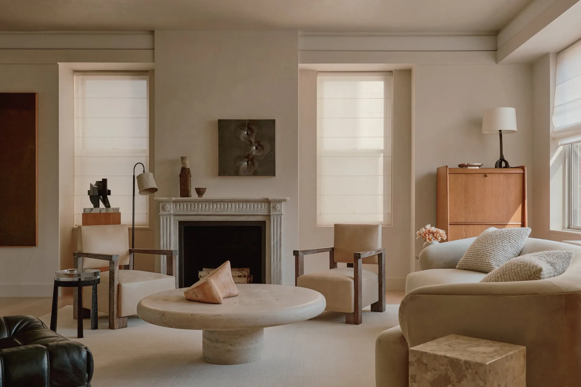 Minimalist living room with neutral palette featuring a cylindrical stone table, beige seating, fireplace, and soft natural lighting by Nicholas Obeid.
