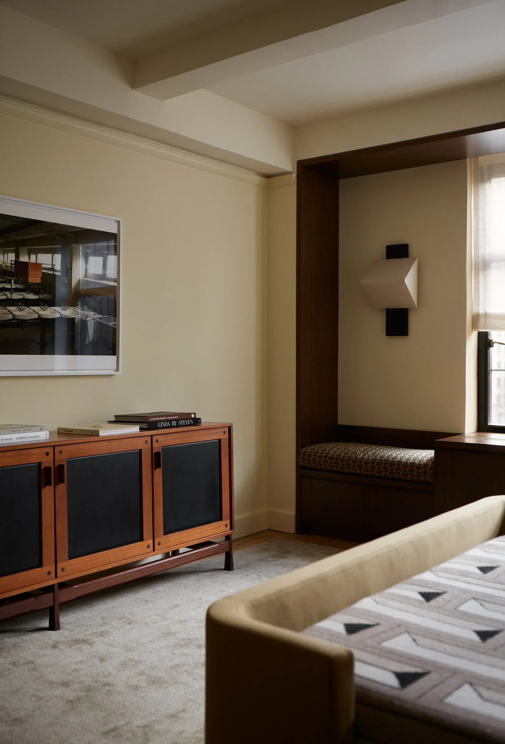 Modern bedroom with warm cream walls, wood-framed bed, orange-and-black dresser, and dark wood architectural elements by Nicholas Obeid.