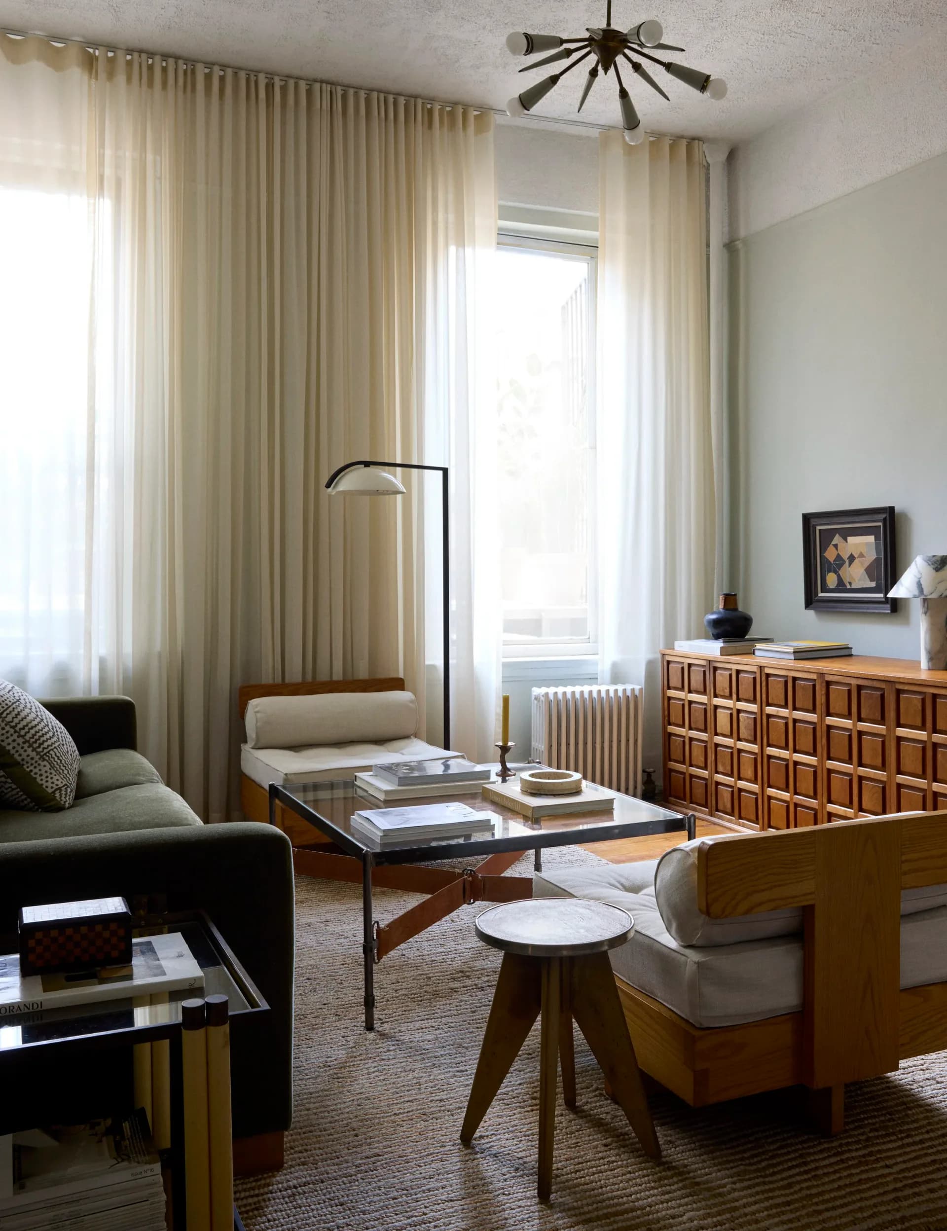 Mid-century modern living room with cream curtains, wooden dresser, metal-frame coffee table, and vintage seating by Nicholas Obeid.