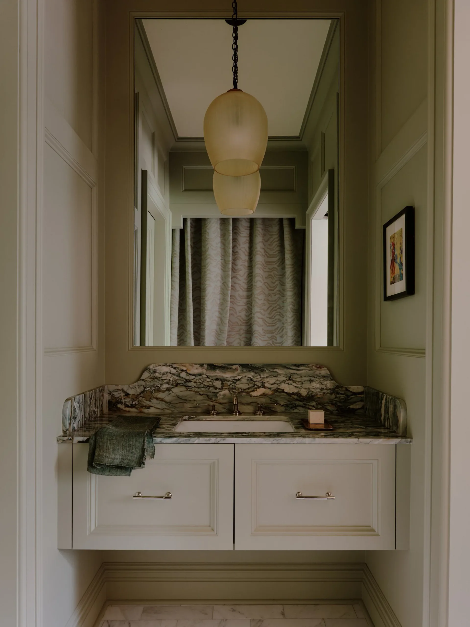 A narrow bathroom vanity alcove with white cabinetry, patterned countertop, round mirror, and hanging pendant light fixture designed by Nicholas Obeid.