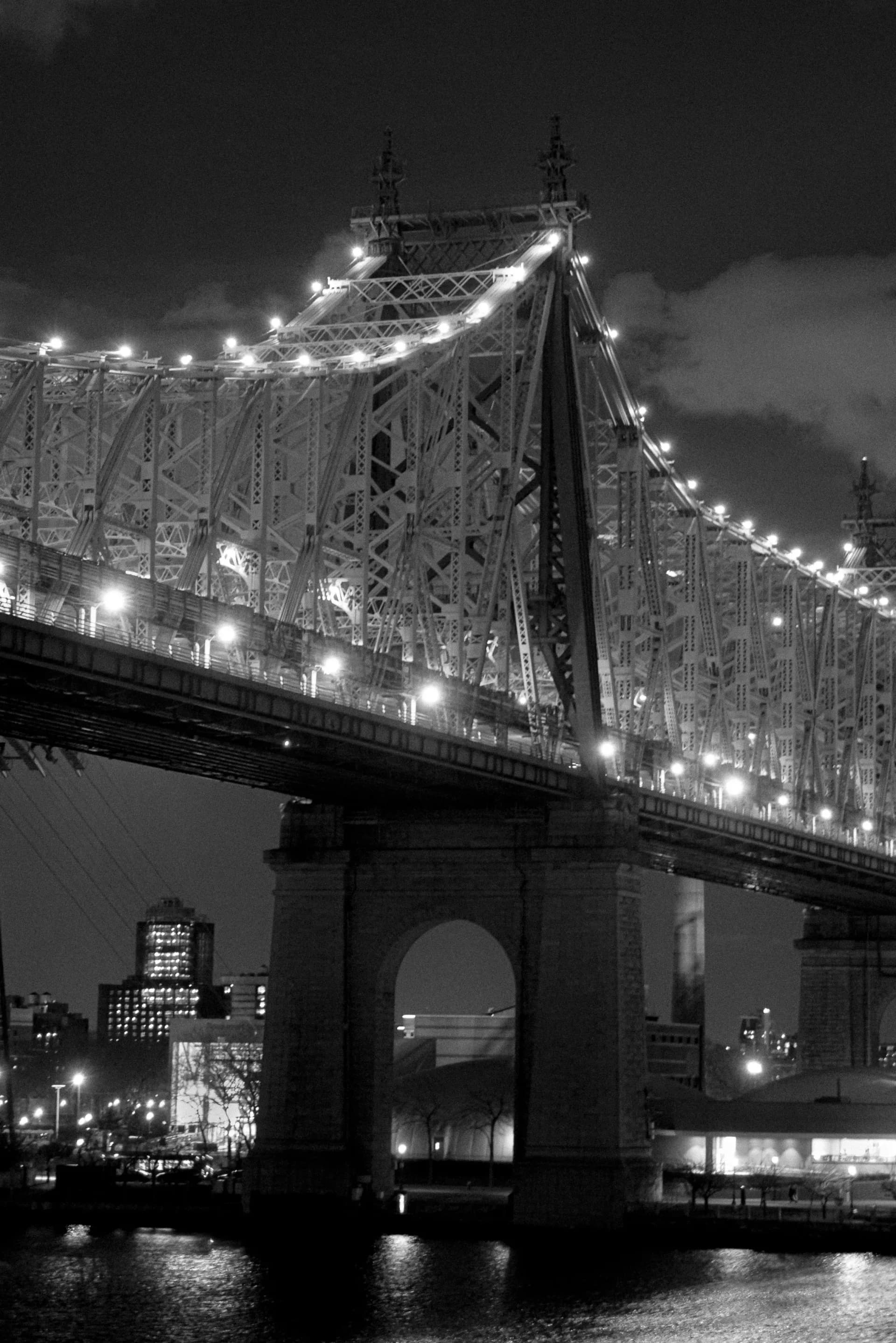 Nighttime architectural photograph of an illuminated steel suspension bridge with ornamental towers, dramatic lighting along cables and deck, dark sky and water below by Nicholas Talamo (WorksbyNET).