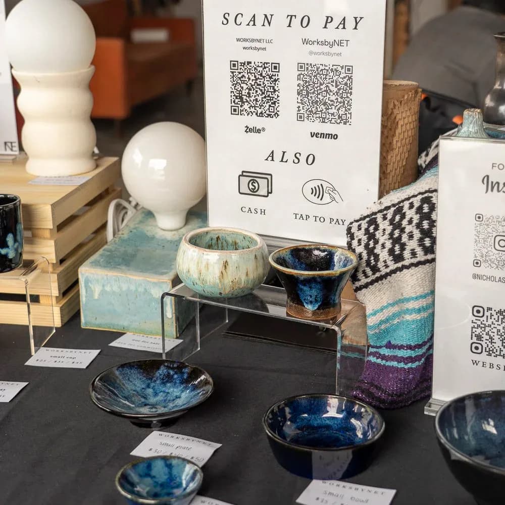 Ceramic tableware display featuring blue-glazed bowls and vessels arranged on black surfaces, with payment QR code signage visible, styled retail or showroom installation by Nicholas Talamo (WorksbyNET).