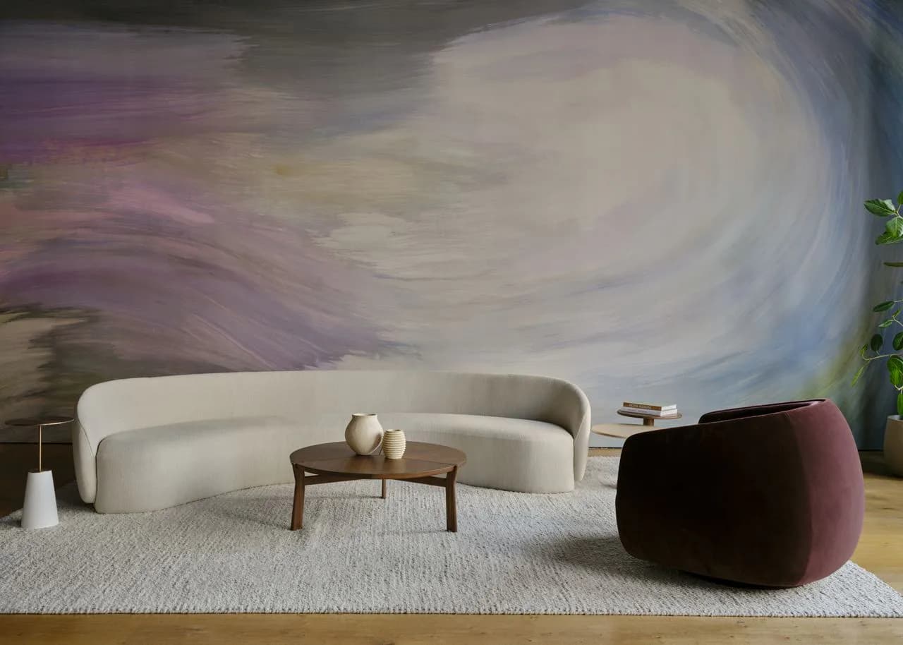 Modern living room with curved cream sofa, burgundy ottoman, and wooden coffee table against abstract purple and white wall mural by Nick Cope.