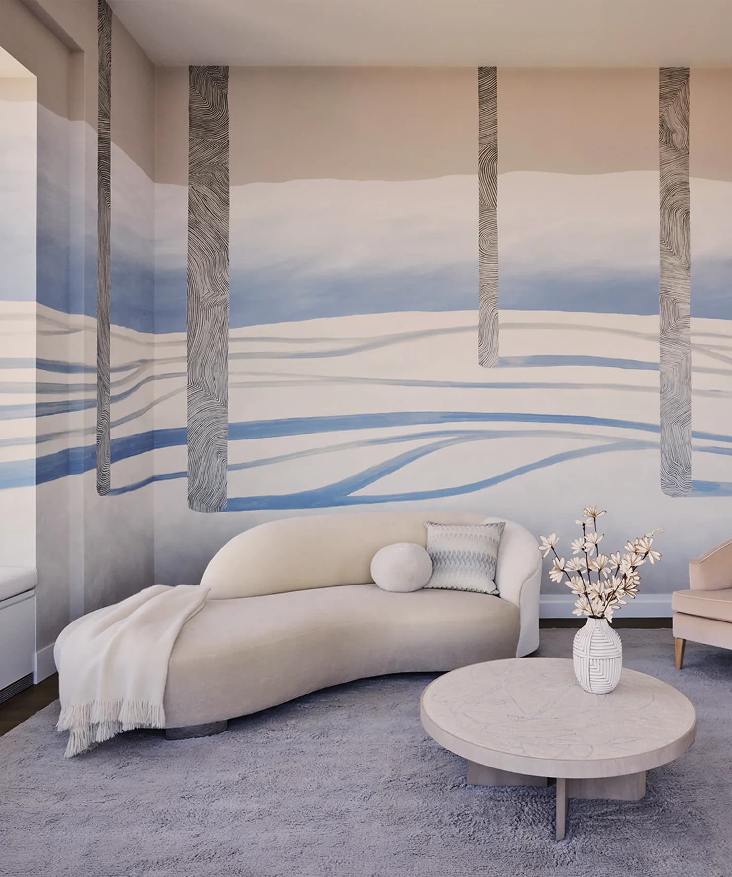 Minimalist bedroom with curved cream sofa, round table, and large-scale mural featuring birch trees and blue wave patterns by Nick Cope.