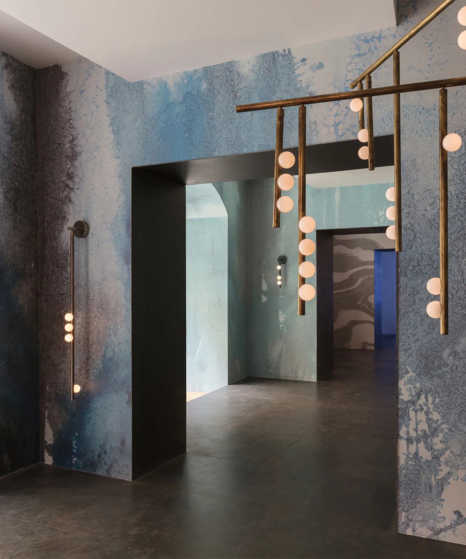 Contemporary interior installation with distressed blue-grey walls, brass-framed mirrors with theatrical lighting, and polished concrete floors by Nick Cope.